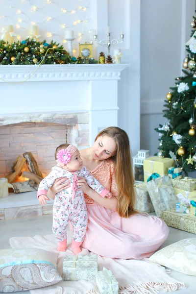 Jovem mãe caucasiana segurando filha nos braços perto de lareira decorada e árvore de Natal, vestindo vestido rosa . — Fotografia de Stock