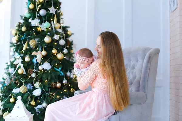 Jovem mulher sentada em cadeira de braço com criança vestindo vestido rosa perto da árvore de Natal . — Fotografia de Stock
