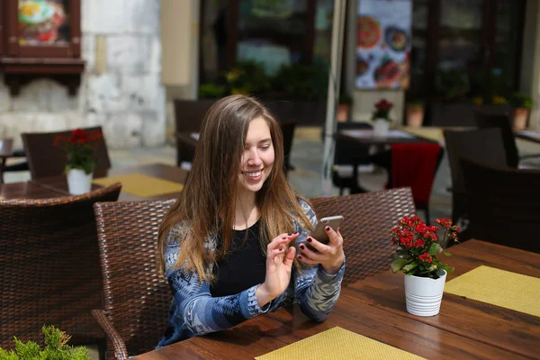 Joven mujer escribiendo mensaje por teléfono inteligente en la cafetería cerca de flores rojas . — Foto de Stock