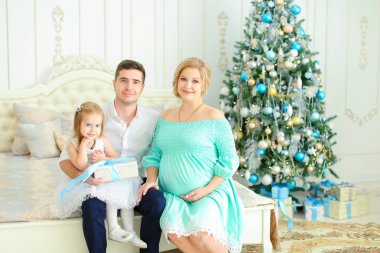 Küçük kızı mutlu baba ve dekore edilmiş Noel ağacı yakınındaki yatakta hamile anne ile oturan.