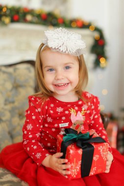 Küçük kız giyiyor kırmızı elbise gülümseyen, hediye tutmak ve dekore edilmiş şömine yakınındaki kanepede oturan.