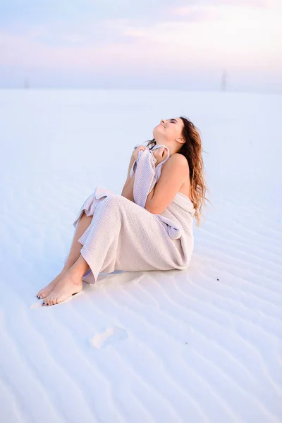 Young woman wrapped in blanket sitting on white sand.