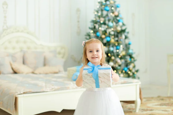 Kleines Mädchen, das im weißen Kleid im Schlafzimmer mit Weihnachtsbaum steht. — Stockfoto