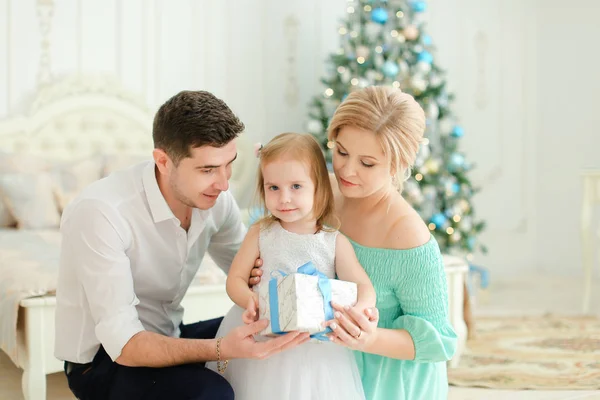 Mujer joven y hombre sentado con hija pequeña manteniendo presente, árbol de Navidad en el fondo . — Foto de Stock