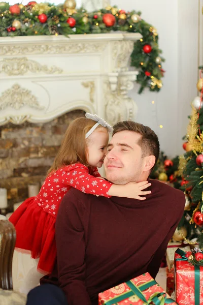 Sonriente padre sentado con su hija pequeña cerca de la chimenea decorada y el árbol de Navidad, manteniendo regalos . — Foto de Stock