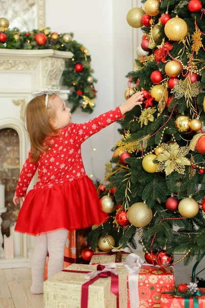 Niña con vestido rojo decorando el árbol de Navidad cerca de la chimenea y regalos . — Foto de Stock