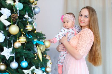Pembe elbise giyen genç beyaz anne holding kız silah ve ayakta yakınındaki hristmas ağaç,.