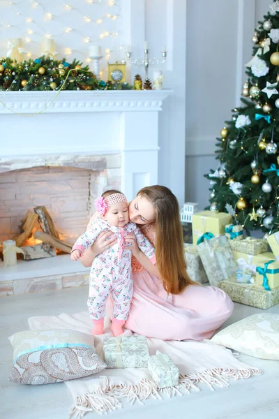 Jovem mãe europeia segurando filha em braços perto de lareira decorada e árvore de Natal, vestindo vestido rosa . — Fotografia de Stock