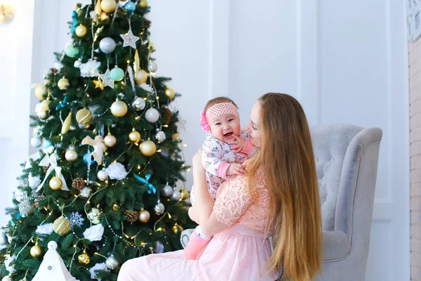 Jovem caucasiana sentada em cadeira de braço com criança vestindo vestido rosa perto da árvore de Natal . — Fotografia de Stock