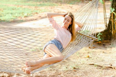 Kot şort giyen kum üzerinde beyaz hasır Hamakta yatan genç sarışın kız.