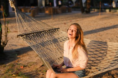 Dizüstü bilgisayar kullanarak ve hasır hamak kuma oturan genç beyaz kız.