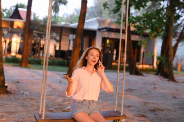 Smartphone ile konuşmak ve oturan sarışın genç kadın salıncak, arka planda kum.