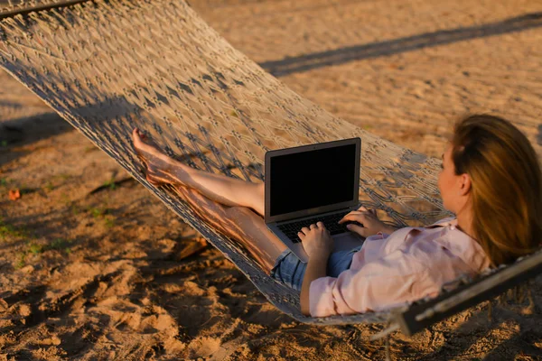 Jovem deitada na rede de vime branco com fundo de areia e usando laptop . — Fotografia de Stock