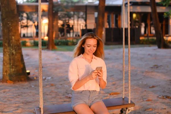 Joven mujer usando teléfono inteligente y sentado en el columpio, arena en el fondo . — Foto de Stock