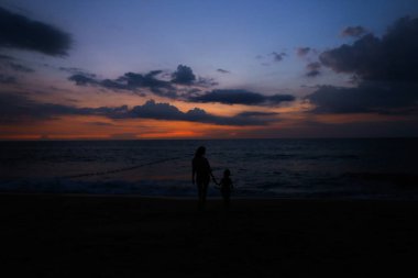 Siyah kızın elini tutarak ve gün batımı artalan ile deniz kıyısında yürüyen Genç Anne silüeti.