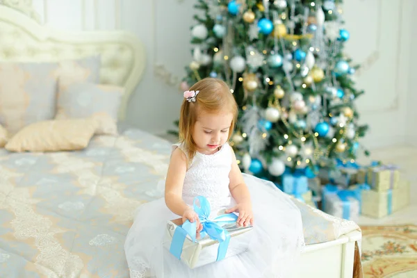 Kleines kaukasisches Mädchen sitzt mit Geschenk auf dem Bett und trägt weißes Kleid, geschmückter Tannenbaum im Hintergrund. — Stockfoto