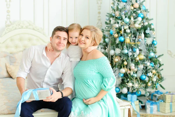 Pequena filha loira sentada com pai feliz e mãe grávida na cama perto da árvore de Natal decorada . — Fotografia de Stock