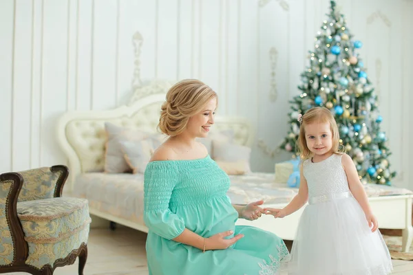 Schwangere glückliche Frau trägt blaues Kleid, umarmt Bauch und sitzt mit kleiner Tochter neben Weihnachtsbaum im Schlafzimmer. — Stockfoto