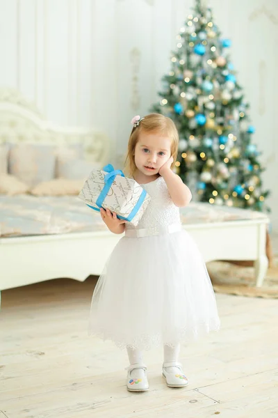 Kleines nettes Mädchen hält Geschenk, trägt weißes Kleid und steht im Schlafzimmer mit Weihnachtsbaum. — Stockfoto