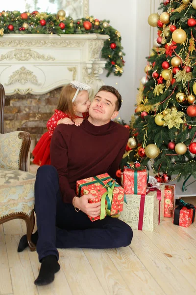 Jovem pai sentado com a pequena filha perto de lareira decorada e árvore de Natal, mantendo presentes . — Fotografia de Stock