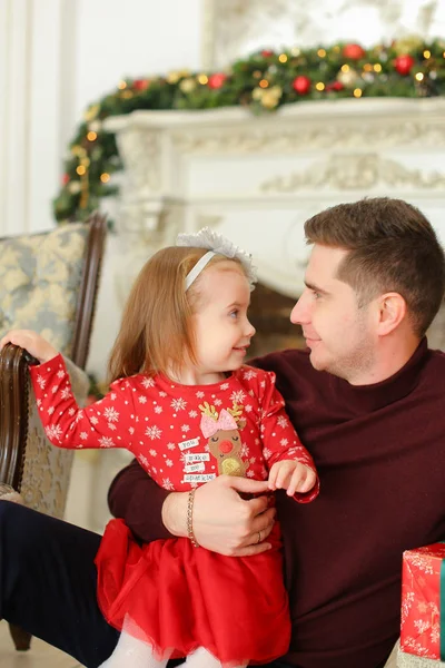 Joven padre sentado con su hija pequeña cerca de la chimenea decorada y manteniendo regalos . — Foto de Stock