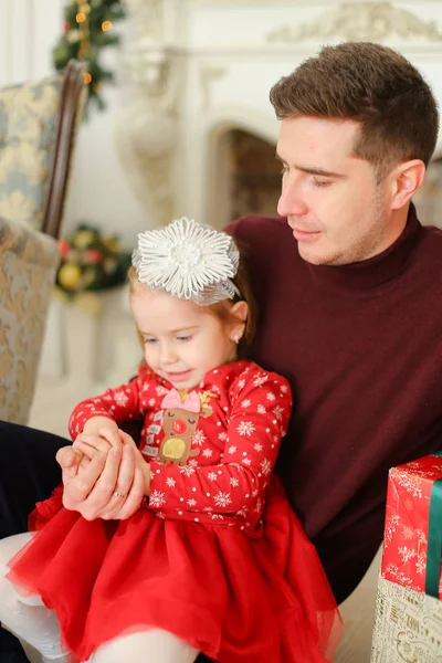 Pequena filha loira vestindo vestido vermelho sentado com o pai perto da parede decorada . — Fotografia de Stock