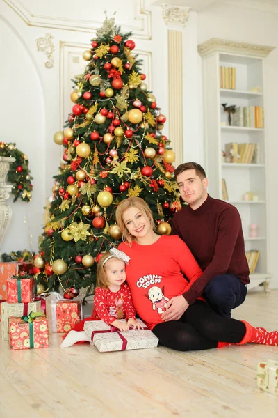 Pequena filha agradável sentada com pai e mãe grávida perto da árvore de Natal e mantendo presentes . — Fotografia de Stock