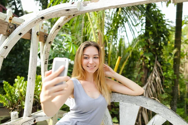 Young caucasian girl making selfie by smartphone with palms in background, sitting on swing. — Stock Photo, Image