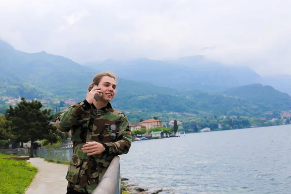 Beszél a smartphone közelében Lépcsőkorlát, a Comói-tóra és az Alpok hegyi háttérben hosszú hajú fiatalember. — Stock Fotó