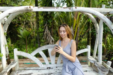 Salıncakta sürme ve egzotik Bahçe, avuç içi arka plan smartphone kullanarak sarışın genç kadın.