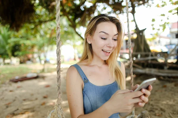 Joven chica bonita utilizando el teléfono inteligente y disfrutar de las redes sociales, montar swing en la arena . — Foto de Stock