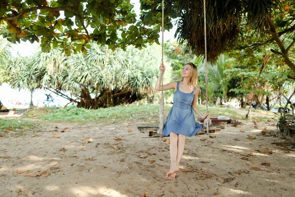 Jonge vrouwelijke persoon draagt jeans jurk mooi en zand op schommel, rijden in achtergrond. — Stockfoto