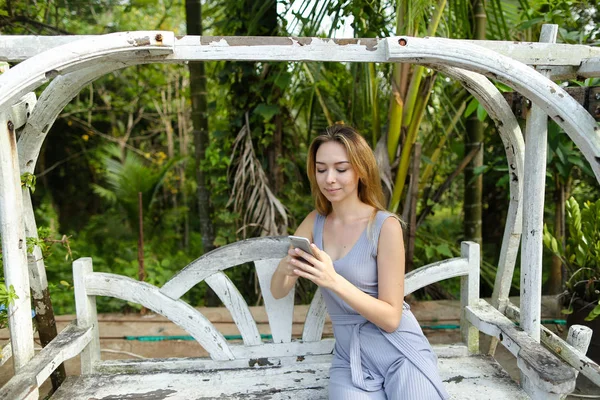 Jovem loira montando no balanço e usando smartphone no jardim exótico, palmas no fundo . — Fotografia de Stock