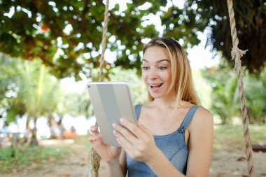 Kot sundress giyen tablet kullanma ve salıncak kum üzerinde sürme genç jocund kadın.