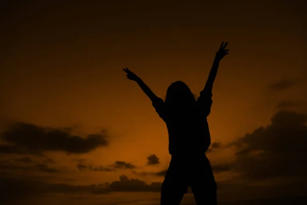 Svart siluett av flicka med upphöjda händer på brun sunset bakgrund med moln i Thailand. — Stockfoto