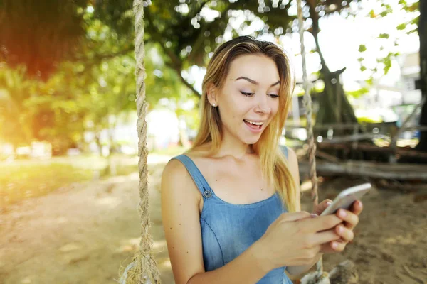 Brillante retrato con sol de chica rubia joven charlando por teléfono inteligente y montar swing en Bali, arena en el fondo . — Foto de Stock