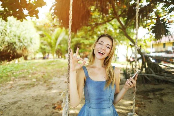 Foto de luz ensolarada de menina montando swing com smartphone na areia e mostrando ok em Bali . — Fotografia de Stock