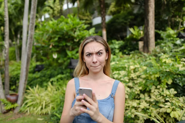Mujer joven haciendo caras y usando sartphone con plantas verdes de fondo . — Foto de Stock