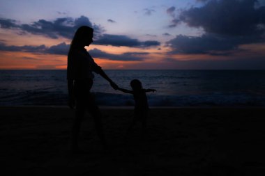 Genç Anne ile küçük kızı deniz kenarında'nde günbatımı yürüme silüeti.