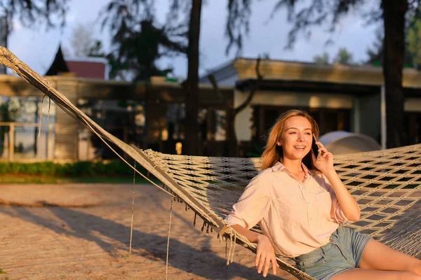 Jovem mulher europeia sentada na rede e conversando por smartphone, férias de verão . — Fotografia de Stock