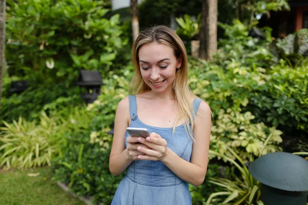 Joven mujer americana charlando por teléfono inteligente en el jardín cerca de los árboles, usando pantalones vaqueros vestido . — Foto de Stock