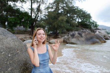 Kum deniz ve taş yakın oturan, smartphone ile konuşmak ve ok gösteren genç aucasian kadın.