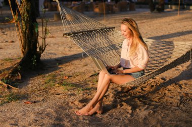 Dizüstü bilgisayar kullanarak ve hasır hamak kuma oturan genç kadın kişi.