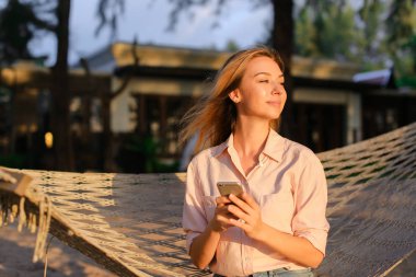 Smartphone kullanarak ve hasır hamak tropikal resort üzerinde oturan genç Avrupa kız.