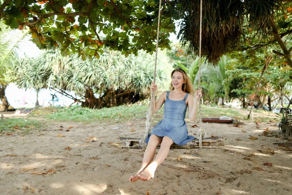 Jonge vrouwelijke persoon dragen jeans jurk en rijden op schommel, zand in de achtergrond. — Stockfoto