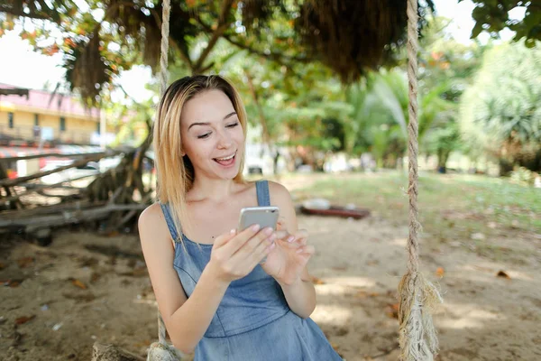 Salıncak sürme ve smatrphone, kum ve ağaç arka planda kullanarak genç kız.