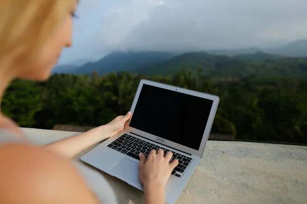 Joven chica bonita que trabaja con el ordenador portátil en el fondo de las montañas verdes, con camisa gris . — Foto de Stock