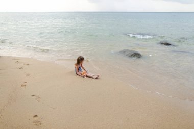 Deniz ve bebekle oynarken yakınındaki kum üzerinde oturan küçük kız.