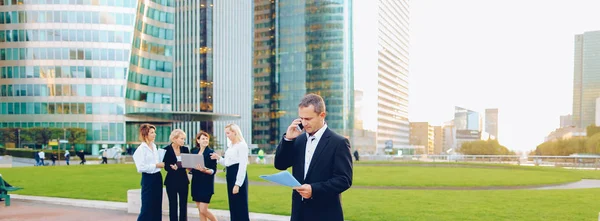 Businessman speaking by smartphone outside, keeping blue document case with employees in background. Concept of boss conversation by mobile phone in La Defense Paris. Business partners talking about