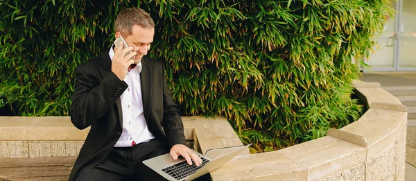 Manager talking by smartphone and working with laptop outside in